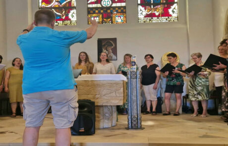 Der Chor steht um den Altar der Kreuzkapelle Gau-Bickelheim und sing. Der Chorleiter steht mit dem Rücken zur Kamera und dirigiert mit gehobenen Armen.